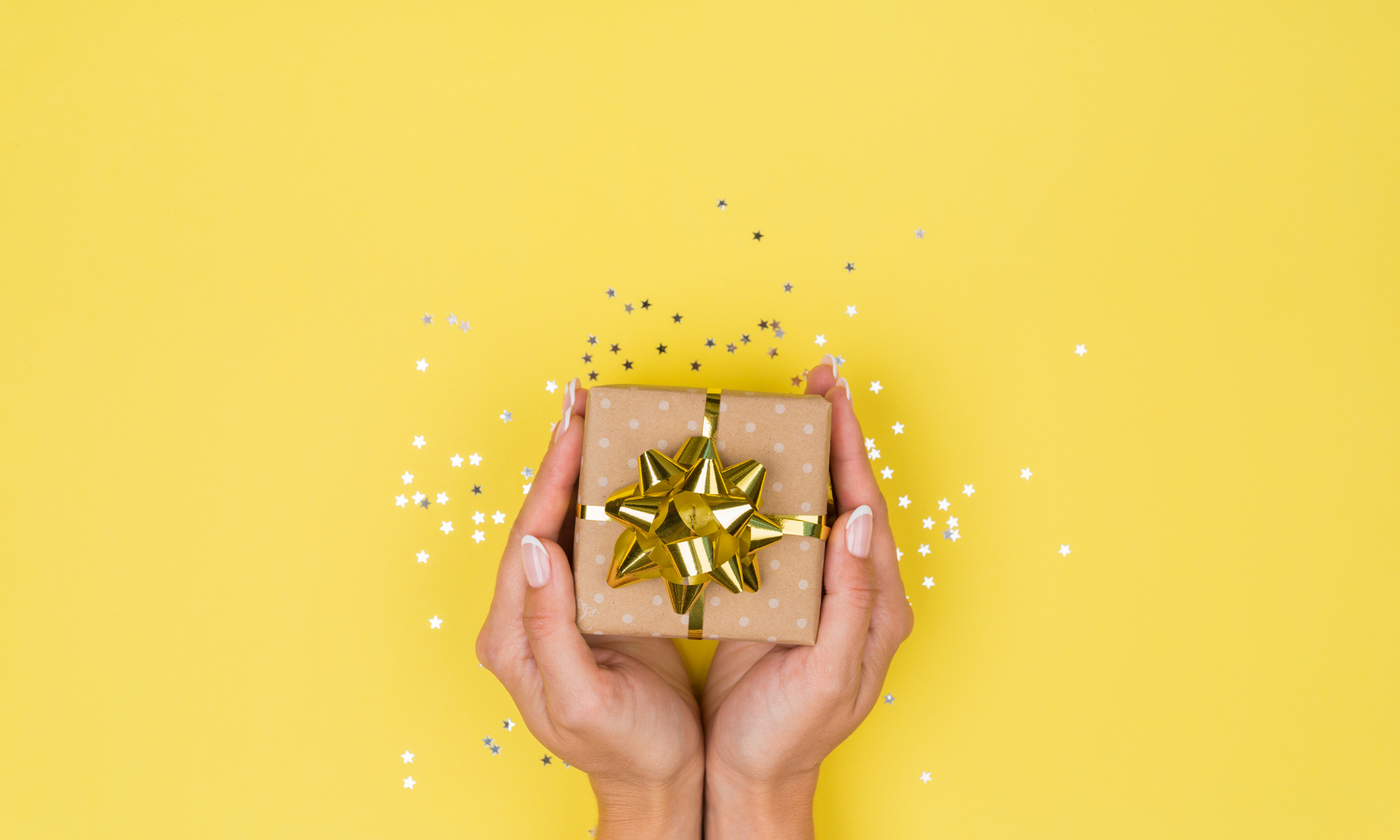 HAND HOLDING GIFT AGAINST YELLOW BACKGROUND, SURROUNDED BY GOLD CONFETTI - THE SWEET REASON CLUB 